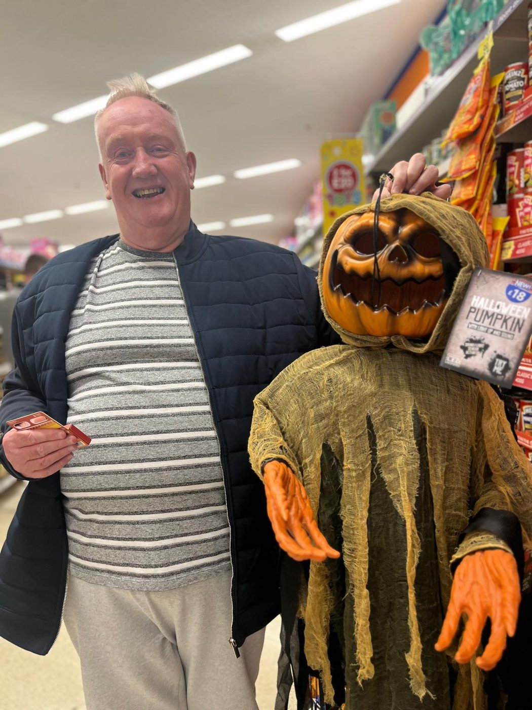 A smiling man in a striped top holding a spooky pumpkin scarecrow figure in a shop.